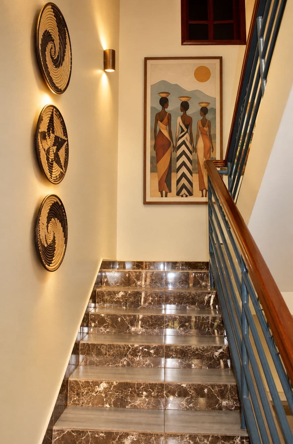 The staircase lit by wall lamps, with woven baskets and a painting of three women