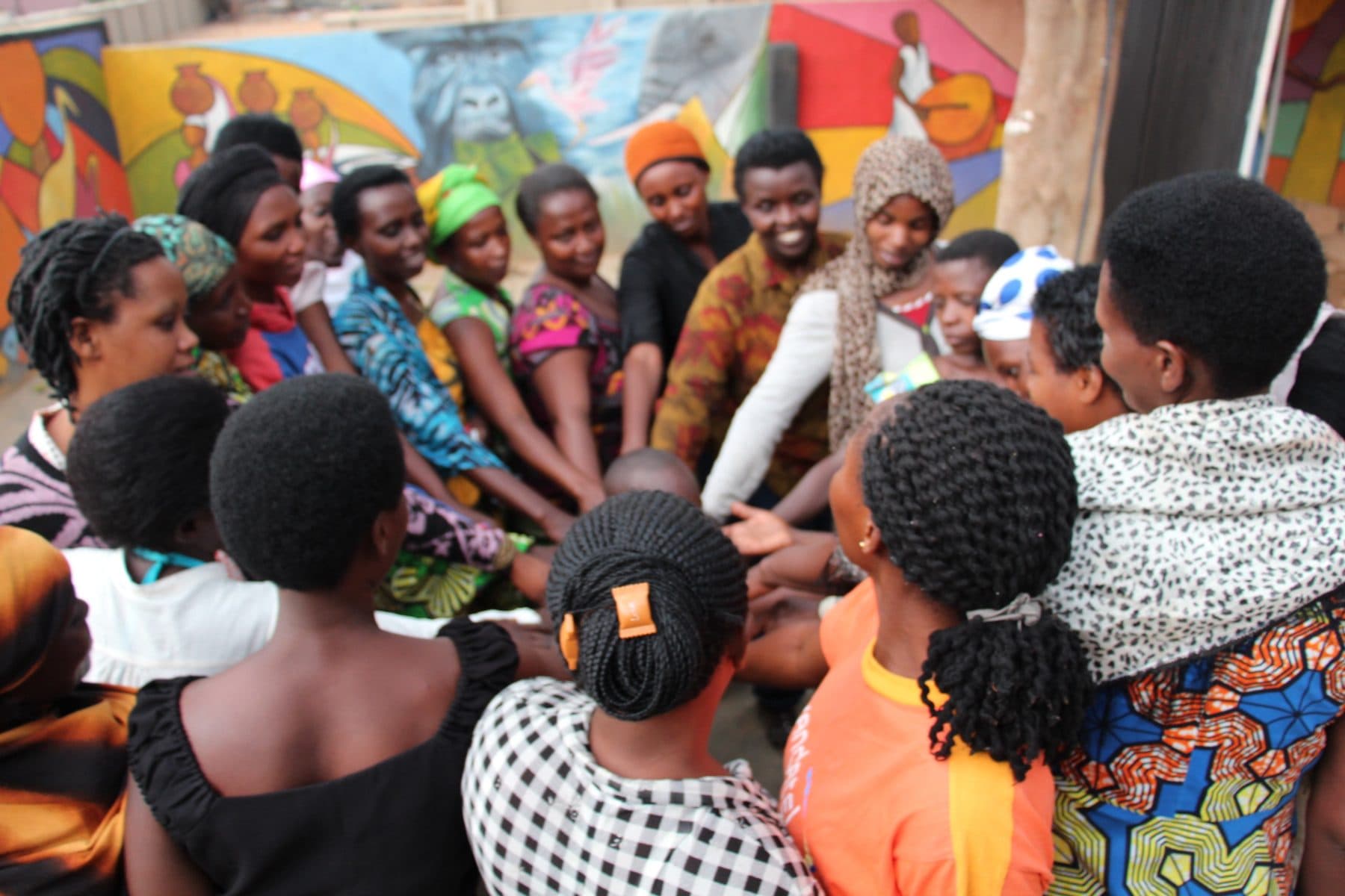 A circle of Mama Rwanda women joining hands in front of a colorful mural